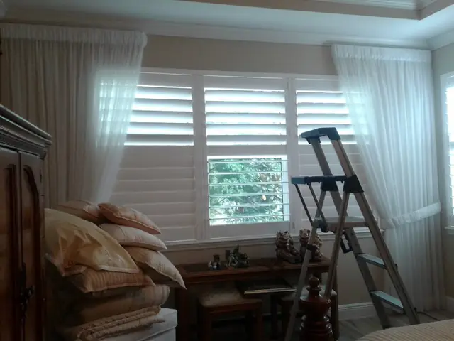 White plantation shutters with sheer side panels in a bedroom window